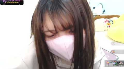 Media: A video of a young Asian woman with long, straight brown hair, wearing a white mask and a white top, sitting on a bed with colorful pillows and a yellow headrest in the background.