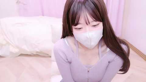 Video of an East Asian woman with long dark hair, wearing a white mask, lavender sweater, and sitting on a white floor, in a soft-pink room with a bed and curtains.