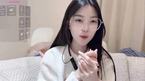 Media: Video of an Asian woman with long black hair, wearing glasses and a white sweater, holding a vape pen. She's on a beige couch in a softly lit room.