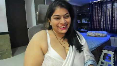 Media: A smiling South Asian woman with long black hair, medium skin tone, and wearing a white blouse with a deep neckline and gold jewelry, sits in a modern kitchen with a gray countertop, glass shelves, and floral curtains.