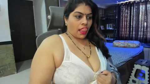 Media: Video of a curvy South Asian woman with medium skin tone, black hair, wearing a white lace bra and gold necklace, licking her lips seductively. Background features a dark room with a bed, curtains, and a chair.