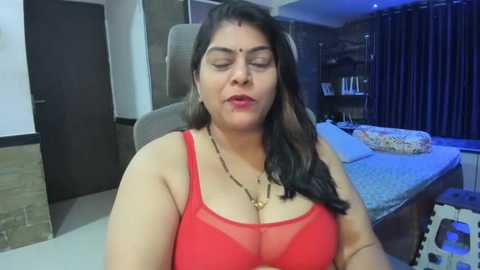 Media: Video of a middle-aged South Asian woman with medium complexion and long dark hair, wearing a red sheer bra, in a modern bedroom with a bed, curtains, and bookshelf.