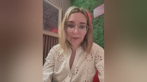 Media: Video of a Caucasian woman with shoulder-length blonde hair, wearing glasses and a white, eyelet-patterned blouse, seated in a room with green artificial grass wall and vintage framed artwork.