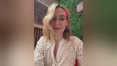 Video of a smiling woman with light blonde hair, wearing glasses, a white lace-up top, and blue earrings, seated outdoors against a green wall and wooden structure.