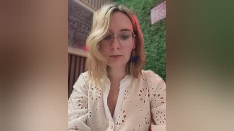 Media: Video of a young Caucasian woman with fair skin and blonde hair, wearing glasses and a white, eyelet-patterned blouse, seated in a lush green environment with a red accent.