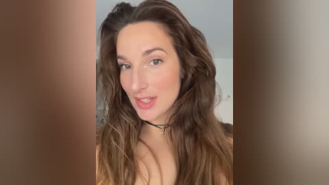 Media: A close-up video of a young Caucasian woman with wavy, medium-length brown hair, fair skin, and full lips, wearing a black choker necklace. The background is blurred, creating a focus on her face.