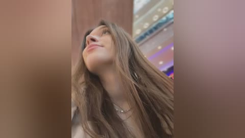 Media: Video of a young woman with long, wavy brown hair, light skin, and a slender build, gazing upward, indoors with blurred background featuring neon lights.