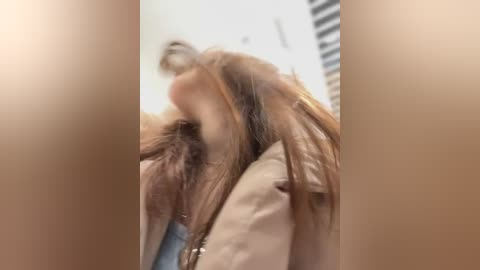 Media: A blurry video showing a person with long, messy brown hair, partially covered in a beige jacket, against a blurred background with horizontal blinds.