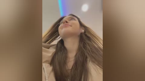 Video of a Caucasian woman with long brown hair, fair skin, wearing a beige top, smiling upwards, blurred background, capturing a serene, happy moment.