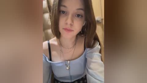 Media: Video of a young woman with light skin and straight brown hair, wearing a light blue off-shoulder top, silver necklace, and black lace skirt. Background shows a beige wall and a door.