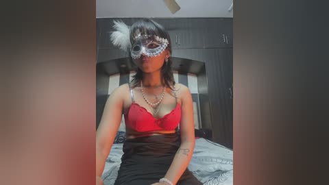 Media: Video of a woman with medium skin tone, dark hair, wearing a red bra, black skirt, and a white feathered Venetian mask, sitting on a bed in a dimly lit room.