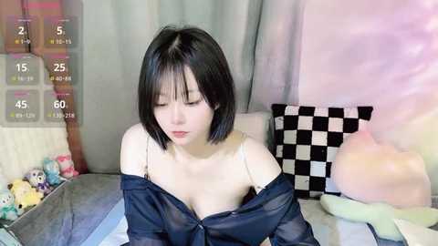 Media: Video of an East Asian woman with short black hair, fair skin, and a slim build, wearing a sheer black blouse that reveals a white bra, sitting on a bed with a checkered pillow.