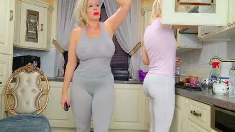 Media: Video of two blonde women in tight gray leggings and tank tops in a luxurious, cream-colored kitchen. One woman with a large bust is adjusting her hair, while the other, also curvy, is bent over the counter.