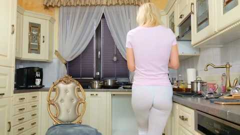 Media: Video of a blonde woman in a light pink top and white pants, standing at a kitchen sink with a view of a window with sheer curtains and a microwave on the left.