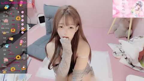 Media: Video of an East Asian woman with long, straight brown hair, wearing a beige off-shoulder sweater, biting her finger, in a pastel pink room with scattered toys and a laptop.