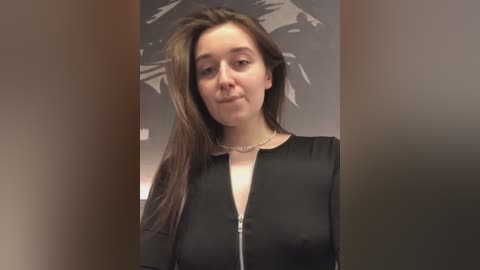 Media: Video of a young Caucasian woman with long brown hair, wearing a black, long-sleeved top with a zipper, and a thin silver necklace. Background features a dark, blurred wall with a mountain pattern. She has a neutral expression.