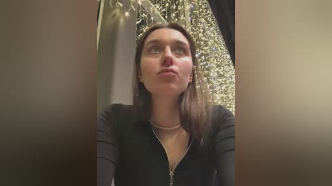 Video of a young Caucasian woman with fair skin, long brown hair, and a slightly pouty expression, wearing a black striped top and pearl necklace, in an indoor setting with a blurred background of greenery and wooden walls.