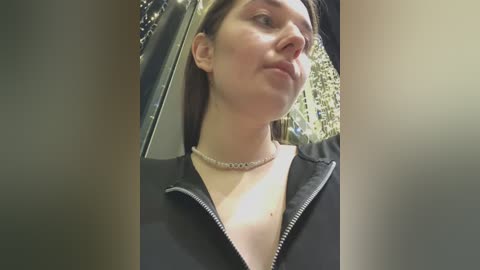 Video of a young woman with fair skin, straight brown hair, and a slender physique. She wears a black zip-up jacket with a silver chain necklace. The background features blurred foliage and a car interior.