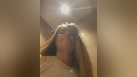 Media: A video of a smiling young woman with long blonde hair, wearing a white shirt, captured from a low angle in a dimly lit room with a ceiling light.