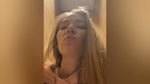 Media: Video of a young woman with long, blonde hair, slightly tousled, making a pouty face, wearing a pink hoodie, in a dimly lit room with wooden walls.