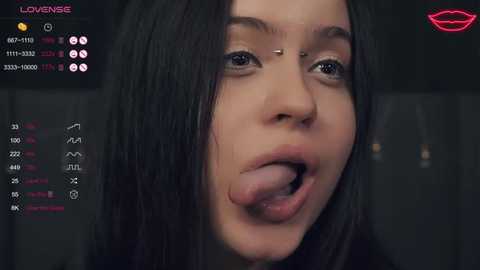 Media: A close-up video of a young woman with long black hair, a nose piercing, and a tongue piercing, sticking her tongue out. The background is dark, with a digital overlay showing her statistics and a lipstick icon.