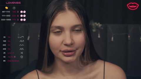 Media: Video of a young woman with long, straight black hair, wearing a black spaghetti strap top, looking slightly bored or tired, in a dimly lit room with a black background and virtual chat interface in the top left corner.