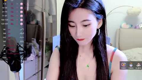 Media: Video of a young East Asian woman with long black hair, wearing a blue dress, looking at a phone screen, in a cozy, cluttered bedroom.