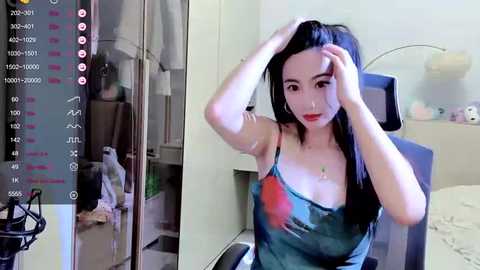 Media: Video of a young Asian woman with long black hair, wearing a blue tank top with a large red heart graphic, sitting in a modern bedroom, adjusting her hair.