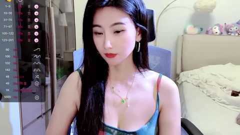 Media: Video of an East Asian woman with long black hair, wearing a low-cut blue top, sitting in a cluttered bedroom with a messy bed, shelves, and a TV.