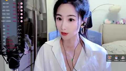 Media: Video of an East Asian woman with dark hair in a ponytail, wearing a white shirt and red lipstick, sitting in a bedroom with a bed and nightstand, captured by a live stream with on-screen overlays.