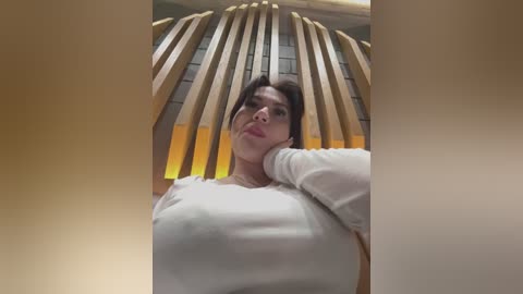 Media: Video of a young woman with medium skin tone, dark hair, and large breasts, wearing a tight white top, leaning against a wooden sculpture in a modern indoor setting.