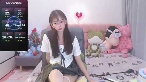 Media: Video of a young Asian woman with straight black hair in a school uniform, sitting on a pink tufted couch in a pastel-themed room with stuffed animals and a digital clock.