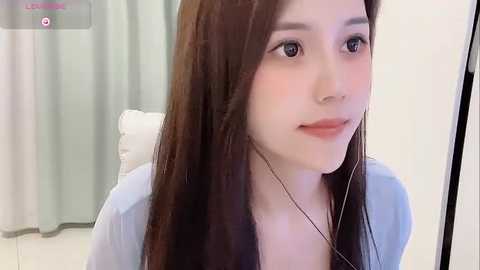 Media: A video of a young East Asian woman with long, straight brown hair, fair skin, and light makeup, wearing a light blue top. She sits in a white room with green curtains in the background.