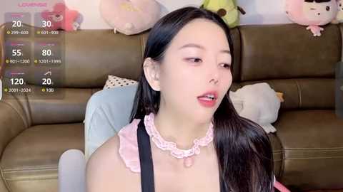 Media: Video of an Asian woman with long black hair, wearing a pink frilly dress and black apron, seated on a brown leather couch. Background includes plush toys and a white pillow.