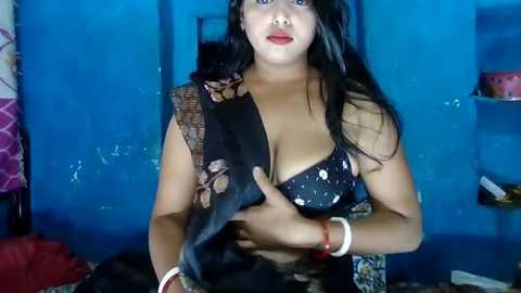 Media: Video of a South Asian woman with long black hair, wearing a black sequined bra, holding a black snake, in a dimly lit, blue-painted room.