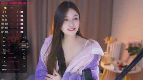 Media: A video of a smiling East Asian woman in a purple, furry robe, indoors with a cozy background and a video game controller.