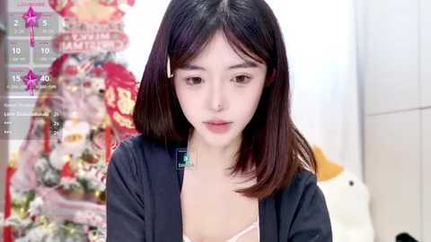 Media: A video of a young East Asian woman with straight, shoulder-length brown hair, wearing a black jacket over a pink top, looking somber in a festive, decorated room.