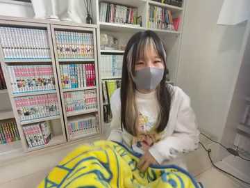 Media: Video of an Asian child with long black hair and a gray face mask, wearing a yellow and blue blanket, sitting on the floor in a brightly lit room filled with colorful children's DVDs and books.