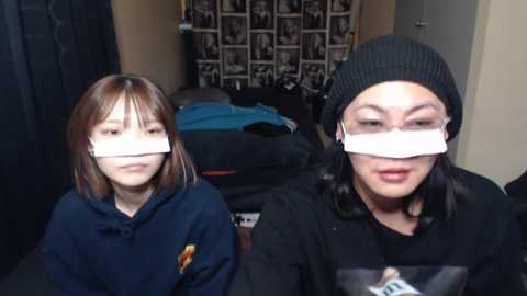 Media: Video of two Asian girls with long hair, wearing black hats and white tape over their mouths, in a messy bedroom with a bed and black curtains.