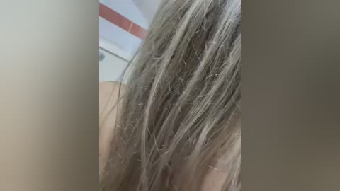 Video of a woman's long, straight, silver-blond hair cascading down her back. She appears to be standing in a bathroom with a tiled background. The image focuses on the texture and length of her hair.