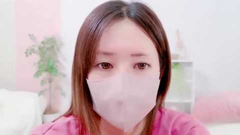 Video of an East Asian woman with straight, shoulder-length brown hair, wearing a white surgical mask, a pink top, and sitting on a white bed in a softly lit, pastel-colored bedroom with a potted plant in the background.