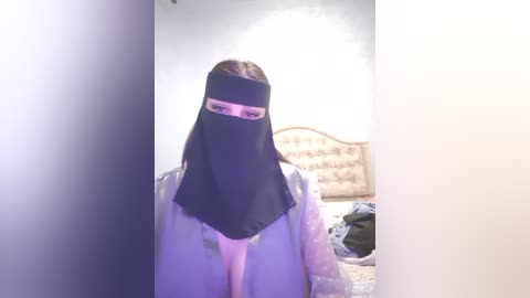 Media: A video of a person wearing a black niqab, standing in a bedroom with a bed, nightstand, and white walls.