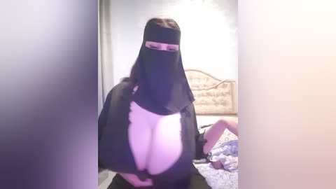 Media: Video of a woman with large breasts wearing a sheer black veil and a revealing black top, standing in a dimly lit bedroom with a bed and decorative wall art in the background.