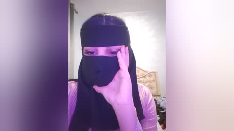 Media: Video of a person wearing a black face mask, looking through a narrow gap, with a blurry background showing a bed and a wall.