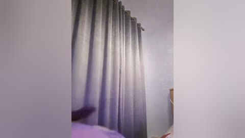 Media: A video of a dimly lit bedroom with soft, lavender-colored curtains drawn to the side, revealing a white wall. A blurry figure, likely a person, is partially visible in the foreground.