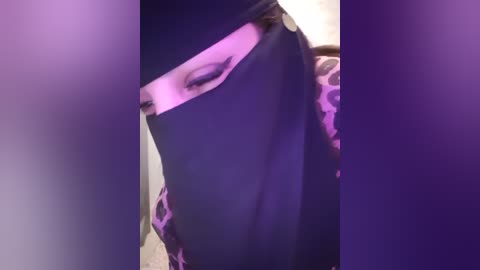 Media: Video of a person with a black mask covering the nose and mouth, wearing a leopard-print shirt. The background is blurred, with purple and white hues. The image has a grainy texture.