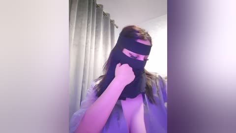 Media: A video of a person with long brown hair, wearing a black mask, holding a hand to their mouth, standing in a bathroom with grey curtains.