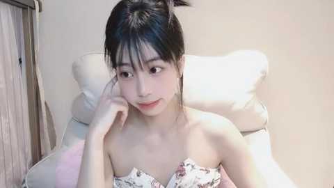 Media: Video of an East Asian woman with straight black hair, wearing a floral off-shoulder top, lying on a white bed in a simple, well-lit room.