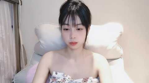 Media: A video of an East Asian woman with fair skin and long black hair tied back, wearing a strapless floral dress, sitting on a white chair with a beige wall and a wooden door in the background.