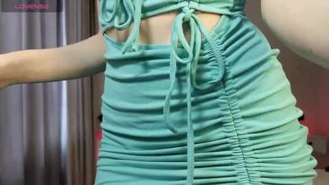 Media: Video of a woman wearing a tight, turquoise, ruched mini-dress with a cut-out design, showcasing her slender waist and small bust, against a blurred indoor background.